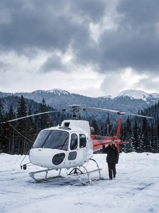 A white and red Airbus AS350 B3 helicopter with three main rotor blades, parked on a packed snow pad under storm cloud, its pilot walking past the tail on a check.