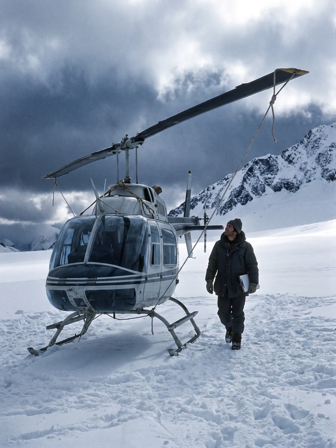 The A-Star tied down on a packed-snow staging pad, the pilot on a walk-around inspection at the tail, storm clouds breaking over the ridge.