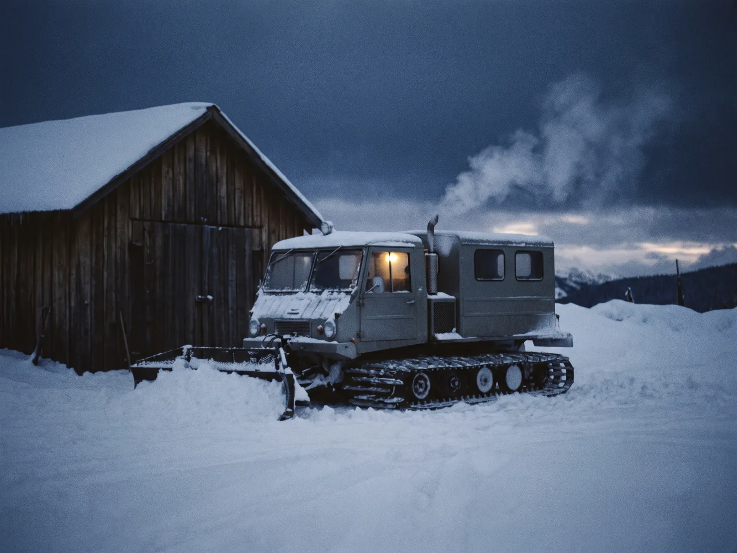A snowcat parked outside the timber machine shed at first light, exhaust steam rising, one warm cab light glowing.