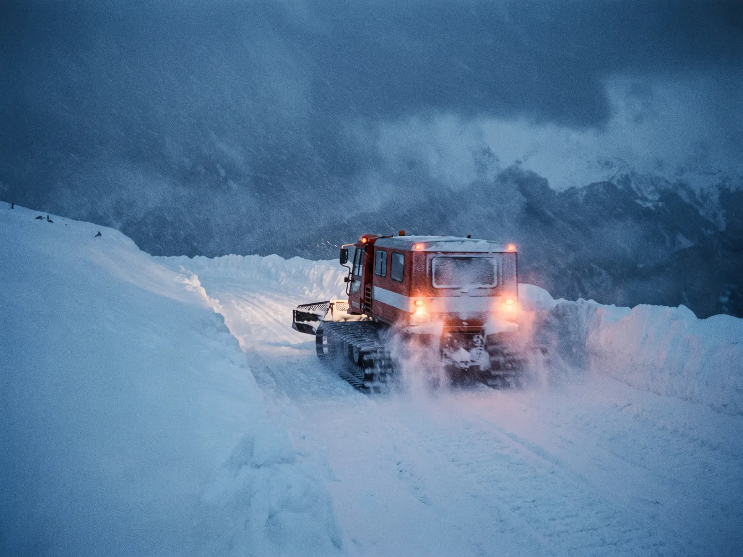 A snowcat climbing a plowed switchback road through deep snowbanks in heavy storm, taillight glow diffused in the flakes.