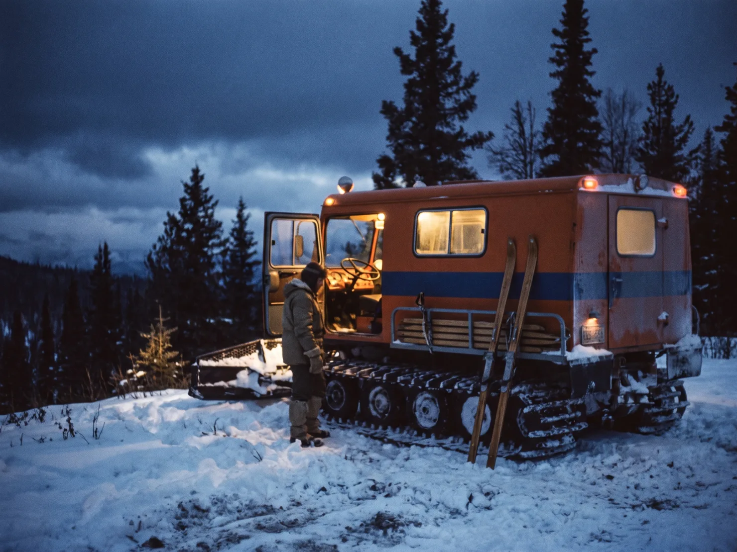A snowcat parked on a treeline bench at blue-hour dusk, cab door open, warm light spilling onto churned snow.