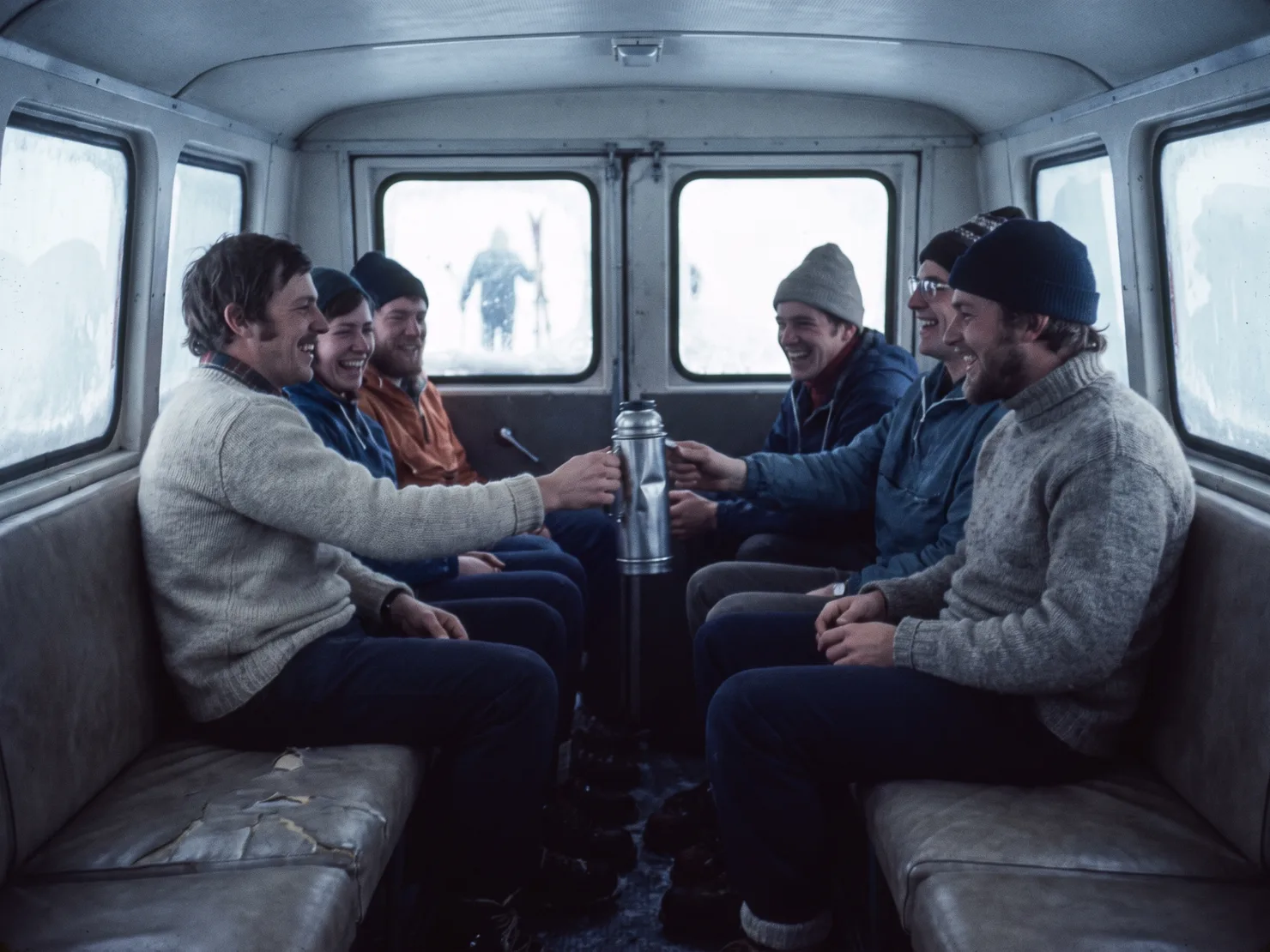 Inside a snowcat's passenger cab: guests in wool sweaters and shell jackets on facing bench seats, laughing, passing a metal thermos, skis standing outside the rear windows.