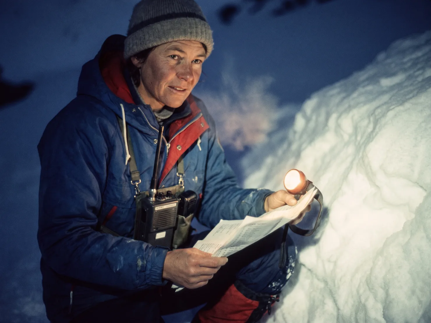 Ren Marchant in a worn guide shell and toque, radio harness on her chest, reviewing the paper run list by headlamp in pre-dawn light.