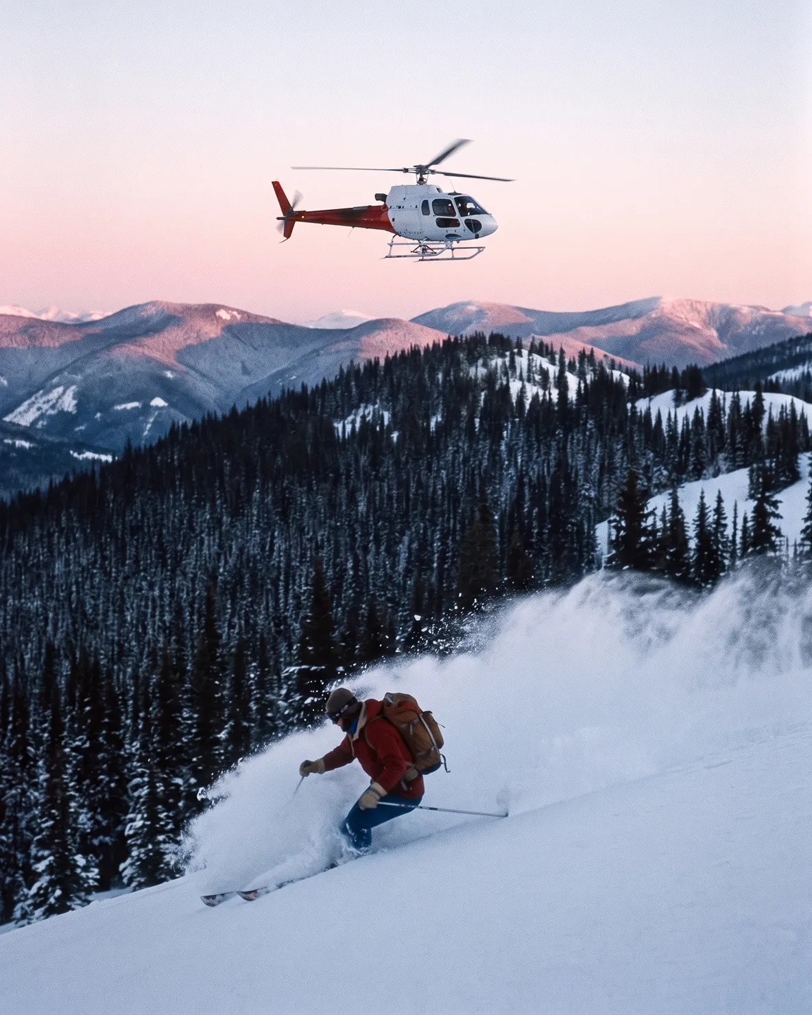 A skier in red wool layers throws a wave of powder mid-turn on a forested slope, while a white and red three-blade helicopter flies above the dark ridgeline in pink pre-dawn light.