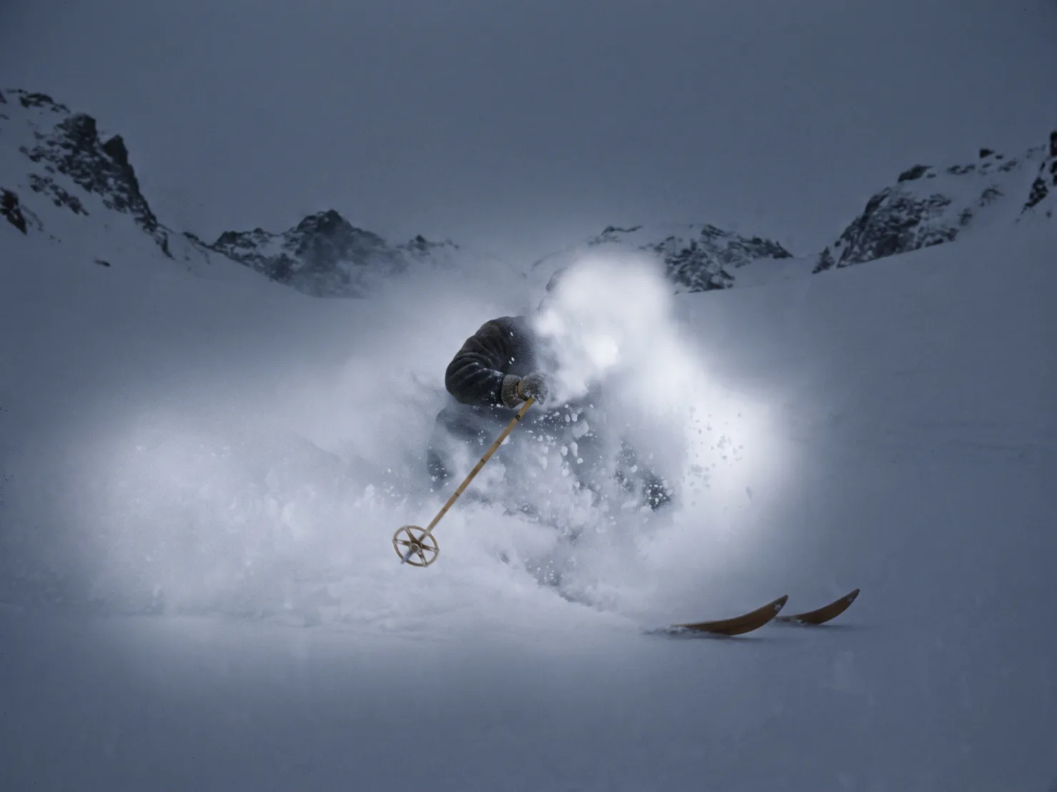 A skier fully inside a cloud of powder at the bowl exit, only a shoulder, one pole and ski tips visible.