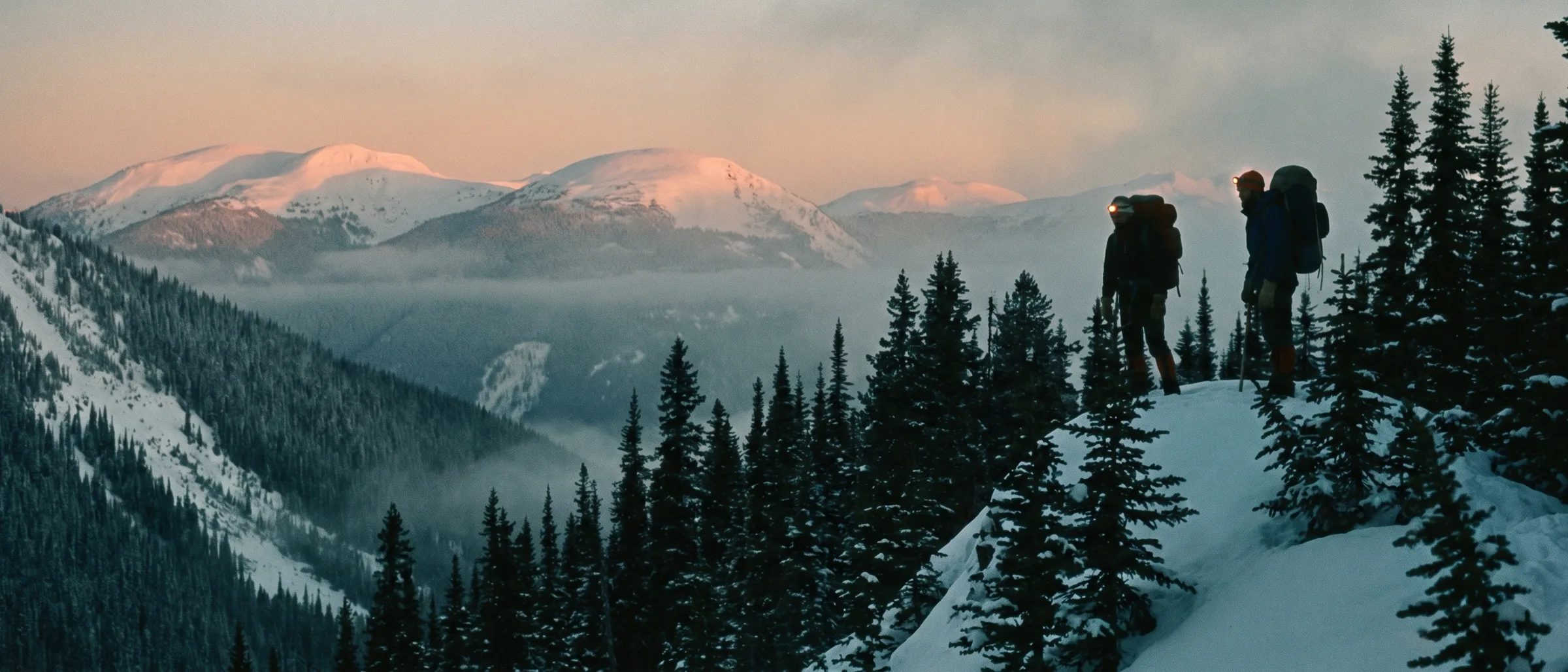 Two guides with packs and headlamps stand on a forested ridge at first light, studying the slope below, rounded summits glowing in the distance.
